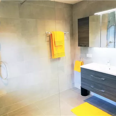 A modern bathroom with a walk-in shower and a sink. Yellow towels and mats provide a cheerful accent.