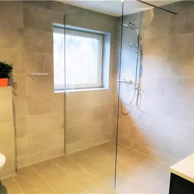 A modern bathroom with a glass shower and a white toilet. There is a window and a small plant container on the windowsill.