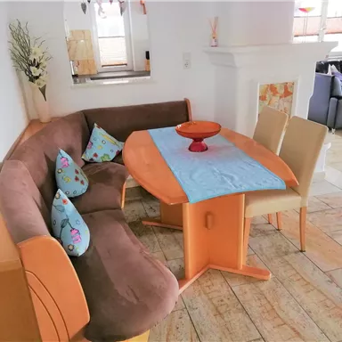 A cozy dining area with a brown corner bench and a table. On the table sits a red bowl and colorful cushions adorn the bench.