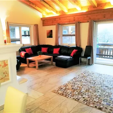 A cozy living room with a black couch and colorful cushions. Large windows offer a view of a balcony and let in plenty of light.