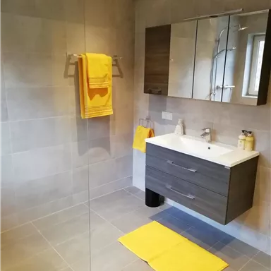 A modern bathroom with gray tiles and a simple sink. Yellow towels and a mat add a colorful accent to the space.