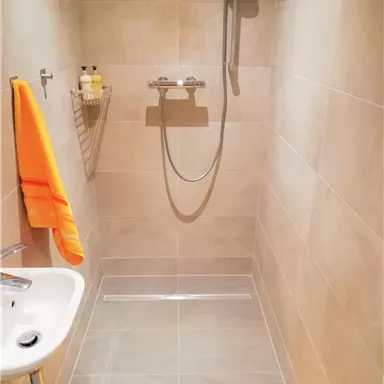 A modern shower room with gray tiles and a hand shower. An orange towel and a yellow bath mat are ready.