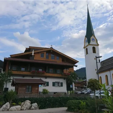 A picturesque village with a traditional church and a wooden house. The sky is partly cloudy and the surroundings are green and inviting.