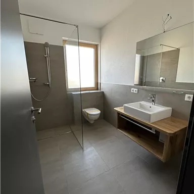 Ein modernes Badezimmer mit Dusche und großem Fenster. Der Waschbeckenbereich hat eine Holzablage und eine elegante Spiegelfläche.