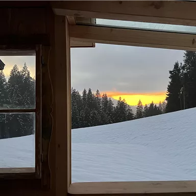 Eine schneebedeckte Landschaft mit Tannenbäumen im Hintergrund. Durch ein Fenster sieht man einen orangefarbenen Sonnenuntergang am Horizont.