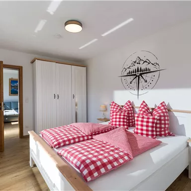 Ein gemütliches Schlafzimmer mit einem großen Bett und karierten Kissen. Der Raum hat einen hellen, modernen Look mit Holzdetails und einem Wanddekor von Bergen.