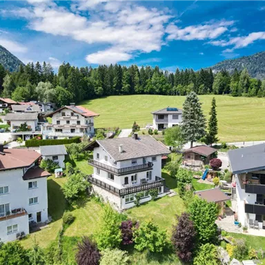 Eine malerische Landschaft mit mehreren Häusern inmitten von Bäumen und Wiesen. Im Hintergrund sind sanfte Hügel und ein blauer Himmel zu sehen.