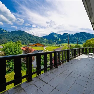 Ein schöner Balkon mit Blick auf die grünen Berge und das blaue Himmel. Im Vordergrund stehen Stühle und die Umgebung ist üppig und einladend.