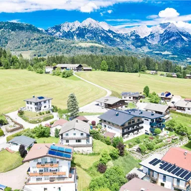 Eine malerische Landschaft mit Häusern in einem grünen Tal. Im Hintergrund erheben sich majestätische Berge unter einem klaren blauen Himmel.