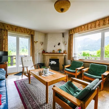 Ein gemütliches Wohnzimmer mit Holzmöbeln und einem großen Fenster, das einen Blick auf die Berge bietet. Der Raum ist hell erleuchtet und mit Pflanzen dekoriert.