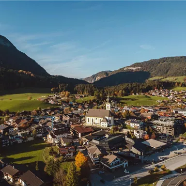 Eine malerische Ansicht eines kleinen Dorfes umgeben von Bergen und grünen Wiesen. Im Zentrum steht eine Kirche, umgeben von traditionellen Häusern und bunten Bäumen.