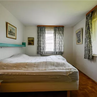 Ein einfaches Schlafzimmer mit einem großen Bett und hölzernem Boden. Vorhänge und Bilder an den Wänden schaffen eine gemütliche Atmosphäre.