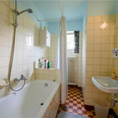 Ein einfaches Badezimmer mit einer Badewanne und einem Handwaschbecken. Die Wände sind gefliest und es gibt einen kleinen Fenster zur Belüftung.