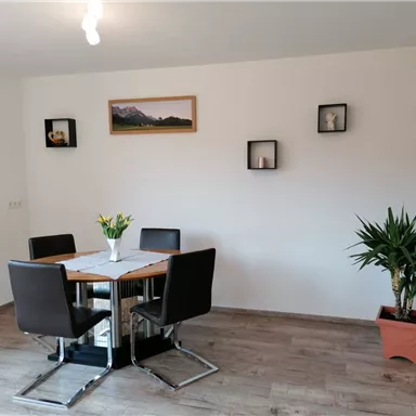 A cozy dining area with a wooden table and modern chairs. Some pictures are hanging on the wall, and a houseplant is in the corner.