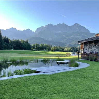 Eine malerische Landschaft mit Bergen im Hintergrund und einem ruhigen Teich. Im Vordergrund sieht man eine grüne Wiese und ein gemütliches Haus.