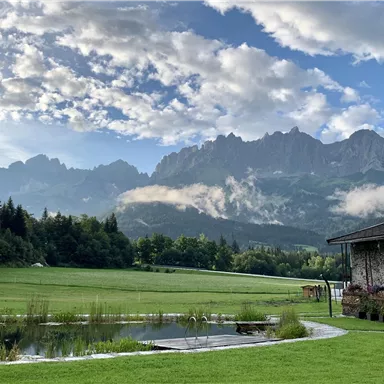 Eine malerische Landschaft mit hohen Bergen im Hintergrund und einer grünen Wiese im Vordergrund. Ein kleiner Teich spiegelt die Wolken und die Umgebung wider.