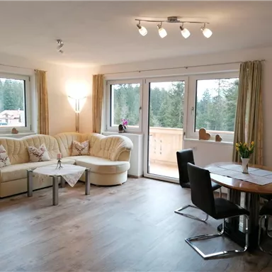 A cozy living room with a bright sofa and a dining table. Large windows offer a view of the nature outside.