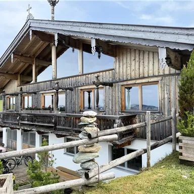A traditional wooden house in the mountains with large windows and a balcony. In the foreground, there are some plants and a pile of stones.