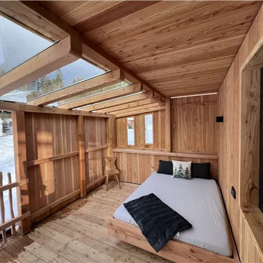 Ein gemütliches Holzhaus mit einem Bett und großen Fenstern. Draußen sieht man Schnee und Bäume.