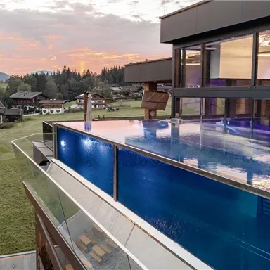 Ein modernes Haus mit einer großen, blauen Infinity-Pool und Blick auf die Berge. Die Umgebung ist grün und ruhig, ideal für entspannende Momente.