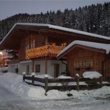 A charming wooden house in the snow, adorned with lights. In the background, snowy trees and a serene winter landscape can be seen.