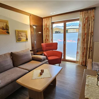 A cozy living room with a gray sofa and a red armchair. Large windows allow a lot of light in and provide a view outside.