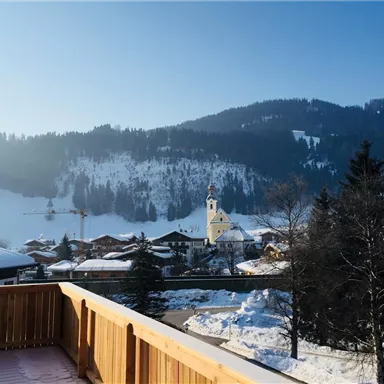 A picturesque winter landscape with snow-covered mountains and a small village. In the foreground, a wooden railing is visible.