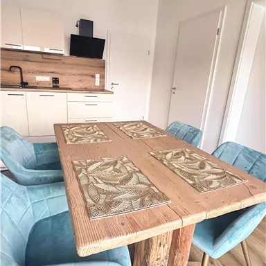 Ein einladender Essbereich mit einem rustikalen Holztisch und blauen Stühlen. Die moderne Küche im Hintergrund ergänzt das schlichte, helle Design.