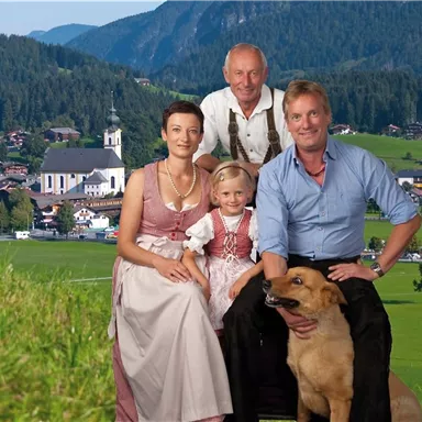A family poses in front of a picturesque mountain backdrop with a dog. They are wearing traditional clothing and smiling at the camera.