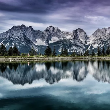An impressive mountain landscape with snow-capped peaks is reflected in the calm water of a lake. The sky is cloudy, giving the scene a dramatic atmosphere.