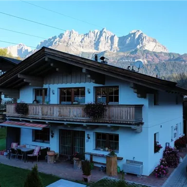 A traditional alpine-style house with a balcony and colorful flowers. In the background, majestic mountains can be seen under a clear sky.