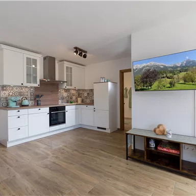 Eine moderne Küche mit weißen Schränken und Holzfußboden. Ein Fernseher an der Wand zeigt eine Landschaft mit Bergen.