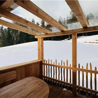 Ein gemütlicher Raum aus Holz mit einem Tisch und Blick auf eine schneebedeckte Landschaft. Im Hintergrund sind Bäume und eine ruhige, verschneite Ebene zu sehen.