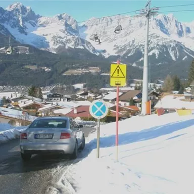 A snow-covered landscape with mountains in the background. A silver car is parked by the road while traffic signs are nearby.