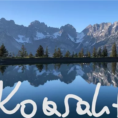 A picturesque mountain landscape with snow-capped peaks and a tranquil lake reflecting the mountains. The sky is clear and blue, making the scene peaceful and inviting.