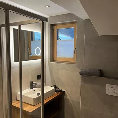 Ein modernes Badezimmer mit einer Glasdusche und einem stilvollen Waschbecken. Die Wände sind in grauem Betonlook gehalten und ein Fenster sorgt für Tageslicht.