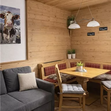 A cozy dining area with wooden paneling and a large table. Next to it is a gray sofa, and two bright lamps hang from the ceiling.