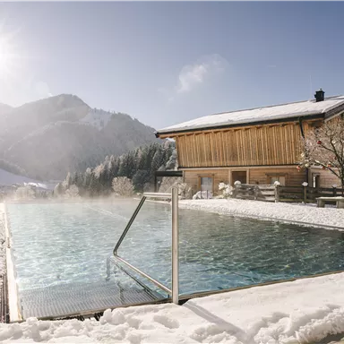 A tranquil outdoor pool in a snow-covered landscape. In the background, a wooden house and surrounding mountains can be seen, while the sun shines.