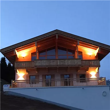 A modern chalet with wooden cladding and a spacious balcony. The lighting creates a cozy atmosphere at dusk.