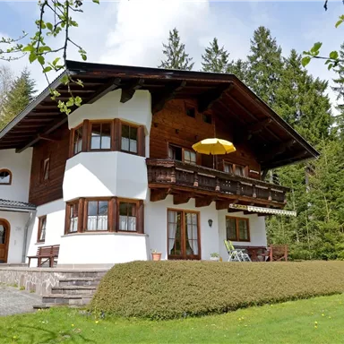 A beautiful chalet in alpine style, surrounded by lush greenery and tall trees. There is a balcony with a sunshade and a well-kept lawn in front of the house.