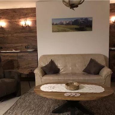 Ein gemütliches Wohnzimmer mit einem beigen Sofa und zwei Sesseln. Der Holztisch in der Mitte ist dekoriert und die Wände sind mit Holzpanelen gestaltet.