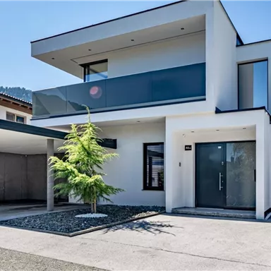 Ein modernes, zweistöckiges Haus mit glatter Fassade und großen Fenstern. Vor dem Haus befindet sich ein kleiner Garten mit einem Baum.