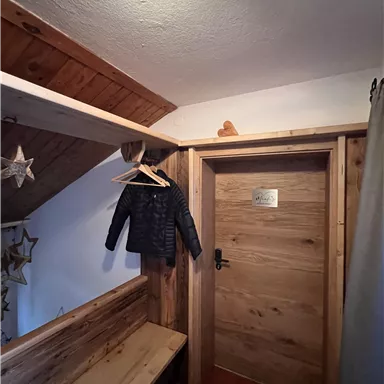 Ein gemütlicher Flur mit einer Holzbank und einem Kleiderhaken. An der Wand hängt eine Jacke, und eine Holztür führt in einen anderen Raum.