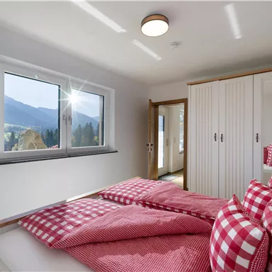 Ein gemütliches Schlafzimmer mit einem großen Fenster, das einen Blick auf die Berge bietet. Das Bett ist mit rot-weiß karierten Kissen und Decken dekoriert.