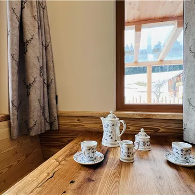 Ein gemütlicher Tisch aus Holz mit einer Teekanne und Tassen aus Porzellan. Durch das Fenster sieht man eine ruhige Landschaft.