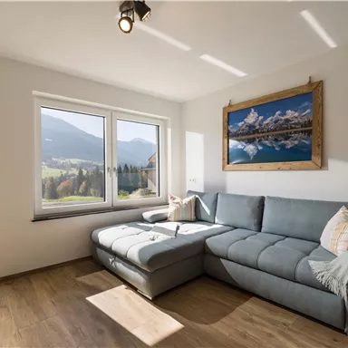 Ein modernes Wohnzimmer mit einem grauen Sofa und einem schönen Blick auf die Berge. Das Licht fällt durch die großen Fenster und schafft eine helle, einladende Atmosphäre.