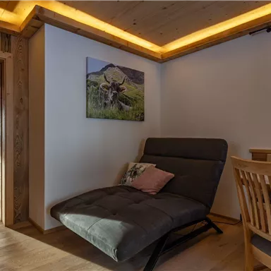 Ein gemütlicher Raum mit einem bequemen Stuhl und einem Holztisch. An der Wand hängt ein Bild einer Landschaft.