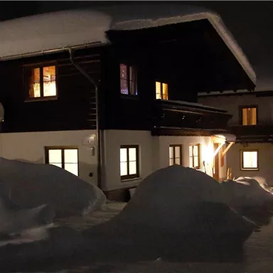 Ein Haus im Schnee bei Nacht. Die Fenster sind beleuchtet und die Umgebung ist von einer dicken Schneedecke bedeckt.