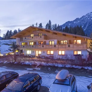 A cozy wooden building in the Alps, surrounded by snow-covered mountains and fir trees. In the foreground, various cars are parked.