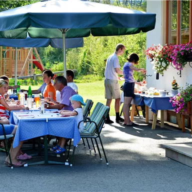 Ein einladender Außenbereich mit einem Tisch, umgeben von Gästen. Im Hintergrund bereitet eine Person ein Buffet vor, während die Sonne scheint.